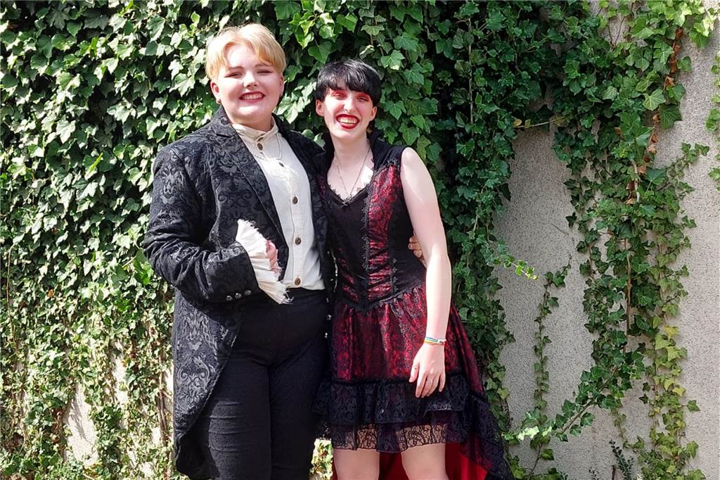 Als Mädchen geboren: Alex Terheyden fühlt sich gefangen im falschen Körper Alex Terheyden und sein Partner Jay Große-Perdekamp grinsen in die Kamera. Alex Terheyden trägt einen schwarzen Mantel mit einem weißen Hemd darunter, sein Partner ein rotes Kleid mit schwarzer Spitze.