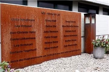 An der Gedenkstätte des Gymnasiums versammelt sich eine kleine Gemeinschaft, um an die beim Flugzeugabsturz am 24. März 2015 getöteten Schülerinnen und Schüler sowie Lehrerinnen zu erinnern. Gesprochene Worte werden in die Klassenräume übertragen.