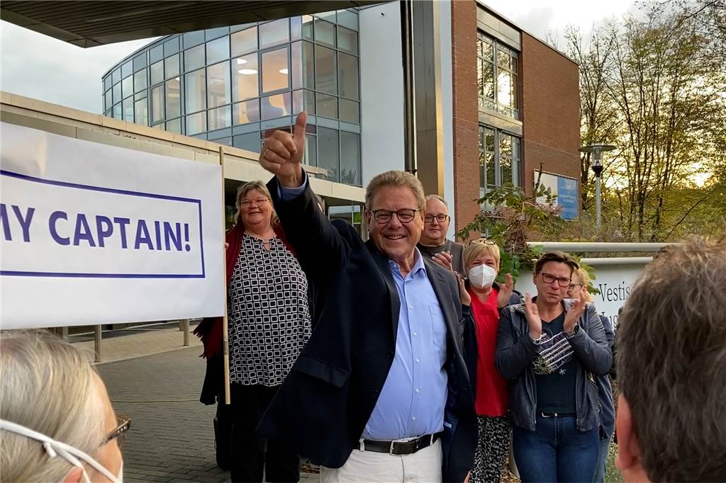 Andreas Wachtel hebt den Daumen seiner rechten Hand. Hinter ihm stehen Krankenhausmitarbeiter, die applaudieren. 