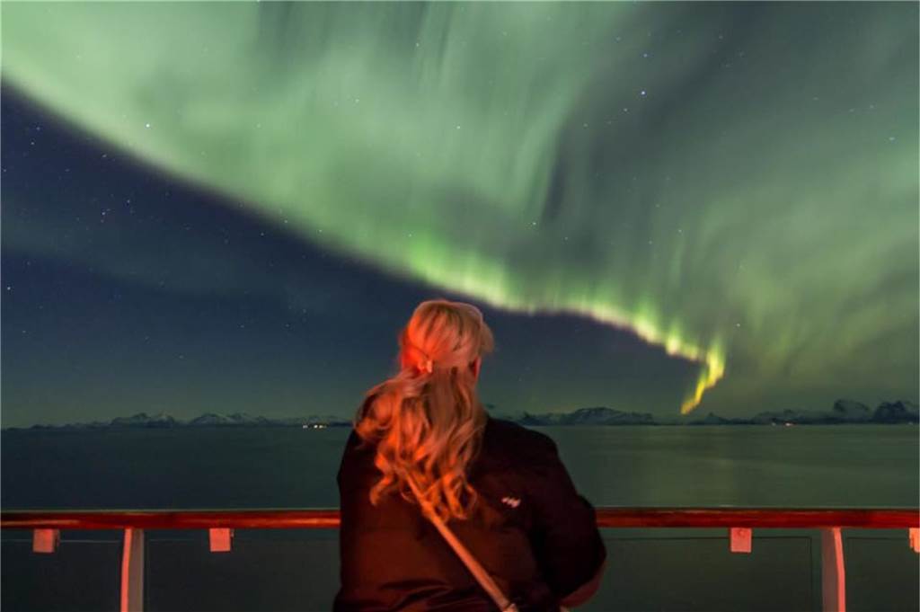 Madleen und Kai flittern unter Polarlichtern Eine Frau steht mit dem Rücken zur Kamera an einer Reling und beobachtet ein imposantes Polarlicht am Himmel.