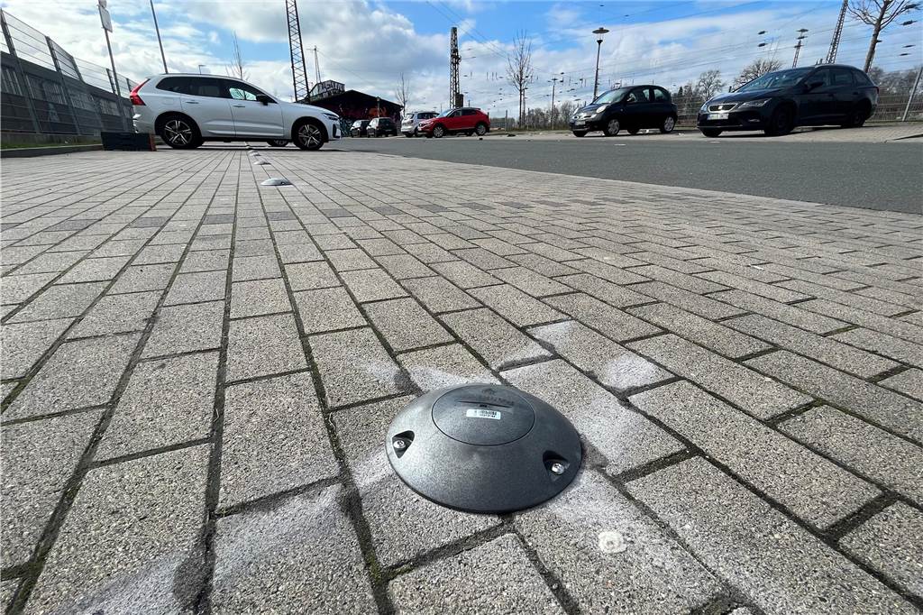 Parkplatz doppelt belegt: Mehr Konsequenzen als teures Knöllchen Aufnahme einer Pflasterfläche mit einem Parksensor. Im Hintergrund sind parkende Autos und Bahnanlagen zu sehen.
