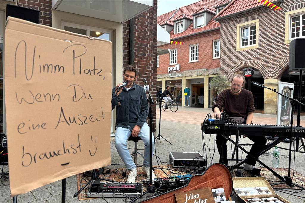 Exklusive Konzerte in Ahauser Innenstadt in der „Sofazone“ Musiker aus Köln machen Musik in Ahauser Innenstadt, davor ein Plakat das zum Platznehmen auffordert