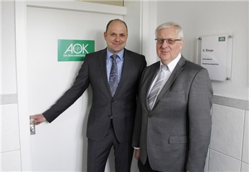 Auf dem ebenfalls beigefügten Foto verlässt Josef Wieteczka (rechts) seinen AOK-Arbeitsplatz und reicht die Verantwortung für die Lüner AOK-Mannschaft an Dirk Staffel (links) weiter.