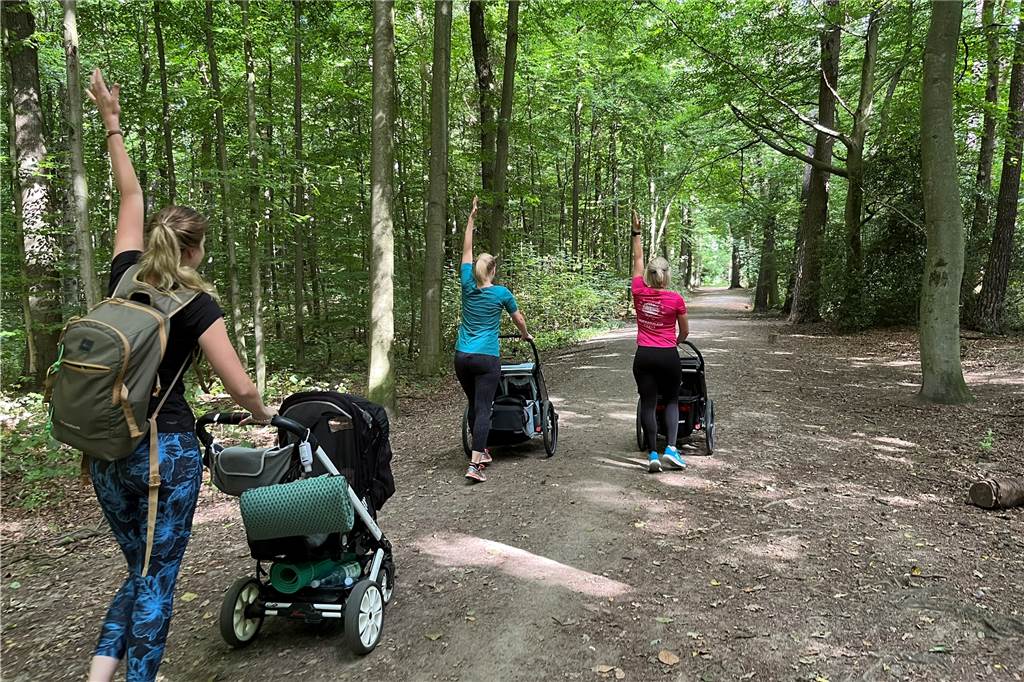Mit dem Kinderwagen durch den Wald: Lisa Steinke (34) bietet Outdoor-Kurse für junge Mütter an Drei Frauen, die ihre Kinderwagen durch den Wald schieben und den linken Arm in die Luft strecken.
