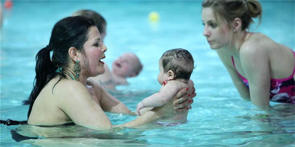 Wassertemperaturen: Babyschwimmen läuft in Castrop-Rauxel weiter Eine Mutter hält ihren Säugling fest und lässt es vorsichtig durch das Wasser eines Schwimmbeckens gleiten.