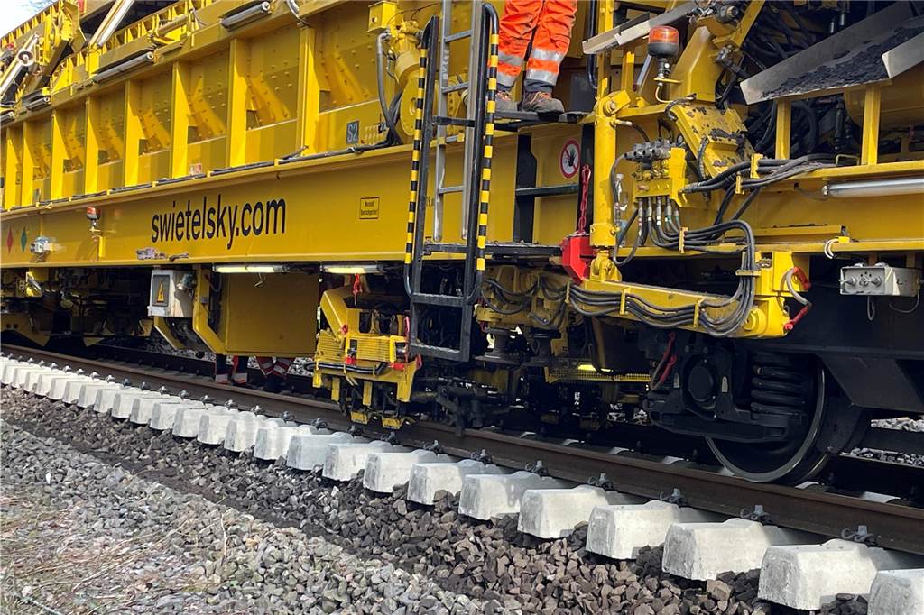 Mega-Maschine tauscht Bahnschwellen und reinigt Schotter im Sekundentakt Bauarbeiter stehen auf dem Zug und sitzen unter dem Zug. Sie überwachen die vollautomatischen Arbeiten der Mega-Maschine. Hier werden die Schienen auf die neuen Beton-Bahnschwellen aufgeschraubt.