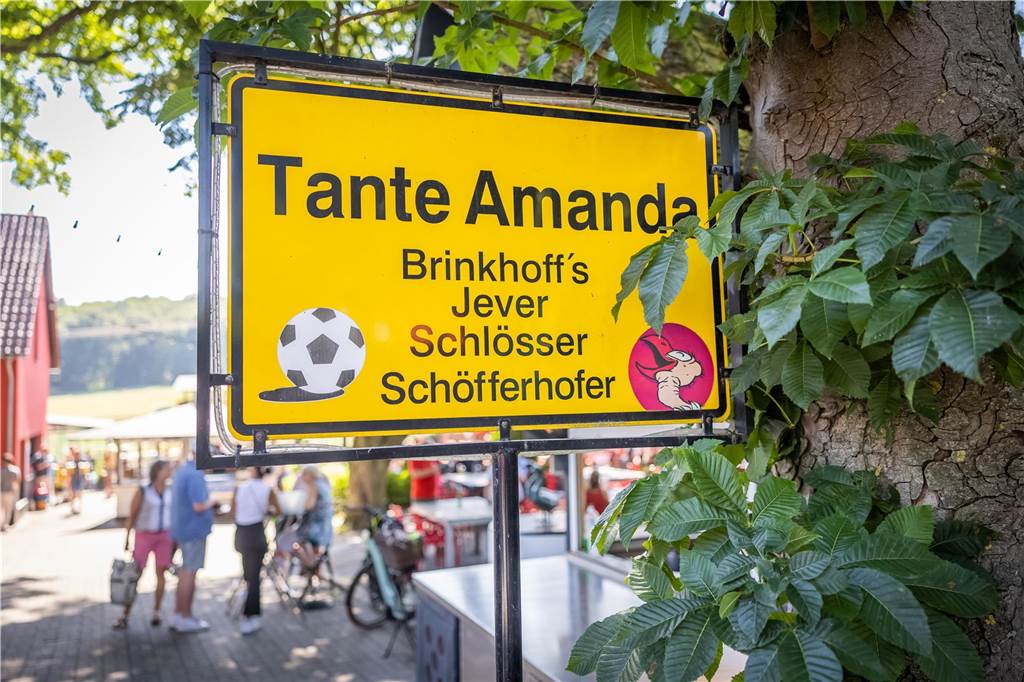 Tante Amanda im Biergarten-Check: Wohlfühloase mit großem Spielplatz Schwarze Schrift auf gelbem Untergrund: Das Schild von Tante Amanda ist einem Ortseingangsschild nachempfunden. Darunter aufgezählt sind die Biersorten, die man hier vom Fass bekommen kann.
