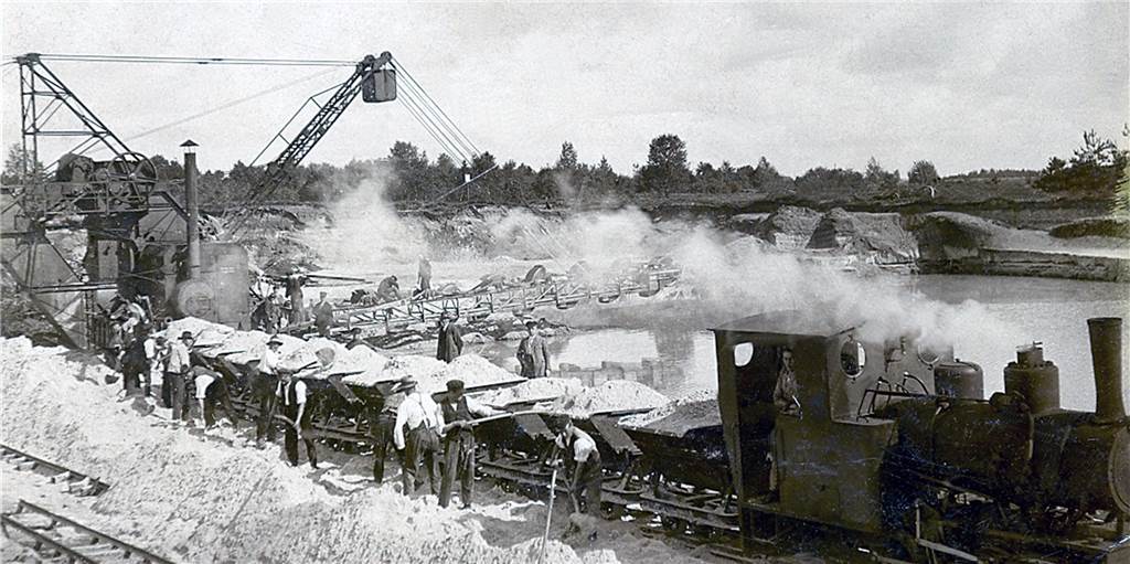 Auf Halterner Sand gebaut: Quarzwerke wachsen beständig Der Grubenbetrieb in Haltern um 1925: Im Hintergrund ist der Gewinnungsteich zu sehen, links ein Dampfeimerkettenbagger. Vorne schaufeln Arbeiter den Sand in die Waggons der Schmalspurbahn.