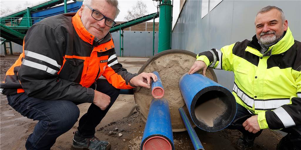 Flüssigboden-Experte Felix Föhre und GWA-Mitarbeiter Thomas Krajewski vor einem Versorgungsrohr, das zu Anschauungszwecken mit dem in Heeren-Werve entwickelten Flüssigboden verfüllt wurde. Der Boden, der sich um diese Leitungen schmiegt, muss nicht verdichtet werden – er verdichtet sich selbst. 