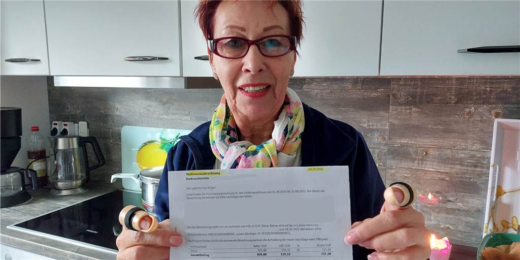 Brigitte Krüger muss die Hälfte ihrer Rente für Heizstrom ausgeben Eine Frau mit roten Haare, einer roten Brille, einem bunten Halstuch und einer blauen Jacke hält eine Heizstromrechnung in die Kamera. Der Gesamtpreis darauf beträgt 721 Euro. Im Hintergrund ist eine Kücheninsel zu sehen. In der rechten Ecke steht eine brennende Kerze.