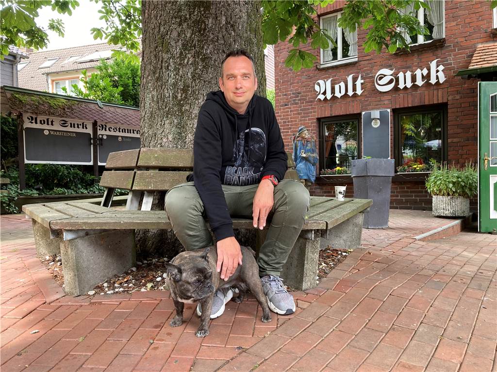 Schlaganfall mit 40: Familienvater kämpft sich zurück ins (Berufs-)Leben Ein Mann sitzt unter einem Baum auf einer Bank und streichelt eine französische Bulldogge, die zwischen seinen Füßen steht. Im Hintergrund ist eine Gaststätte aus rotem Backstein zu sehen. Darauf der Schriftzug "Aolt Surk".