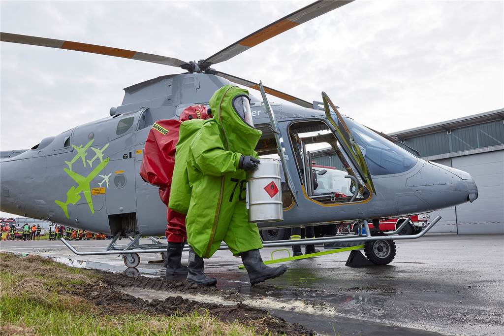 Spezialkraft in neongrünem Schutzanzug trägt weißes Fass mit Gefahrenkennzeichnung von Hubschrauber weg