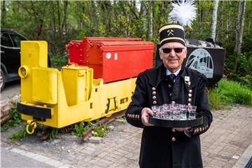 Recklinghausen: Ein Mitglied des Geschichtskreises König Ludwig in Steiger-Montur hält ein Tablett mit Schnapsgläsern in die Kamera. Im Hintergrund ist eine alte Bergbaulok in den Farben Gelb und Rot samt angehängter schwarzer Lore zu sehen.