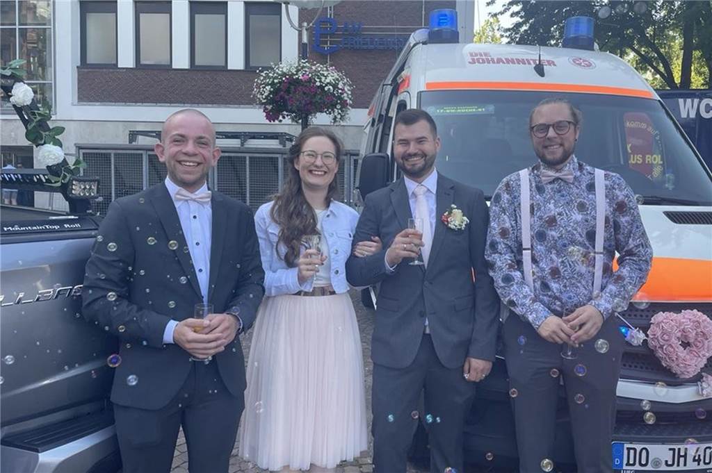 Mit Blaulicht begrüßt: Fabian und Paula haben in Dortmund geheiratet Das Brautpaar steht in der Mitte. Links und rechts von den beiden stehen zwei Männer, die Trauzeugen. Hinter ihnen der mit rosafarbenen Blumen geschmückte Rettungswagen.