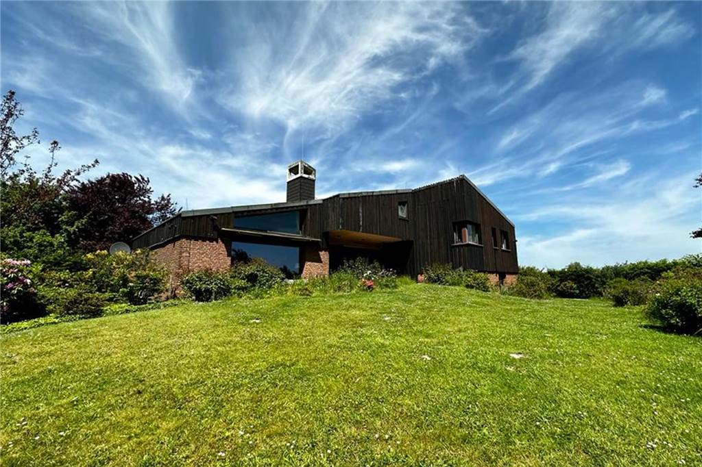 Extravagante Immobilien: Fußbodenheizung im Keller oder hohe Fenster Ein Holz aus Backsteinen im unteren und Holz im oberen Bereich. Im Vordergrund ist eine große Wiese, der Himmel ist blau und die Sonne scheint,