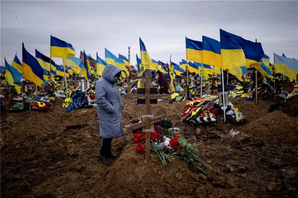 Tote und Verletze bei Raketenangriff auf Cherson: Selenskyj spricht Ukrainern Mut zu Eine Frau steht an einem Grab mit einem Holzkreuz und Blumen. Im Hintergrund sind dutzende Gräber mit ukrainischen Flaggen zu sehen.