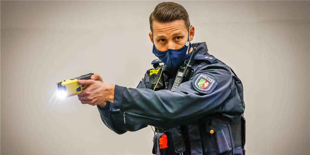 Bodycams können mit Tasern verbunden sein - Polizei darf das aber nicht nutzen Ein Polizist hält einen Elektro-Taser in der Hand und hat eine Bodycam an seiner Schulter befestigt.