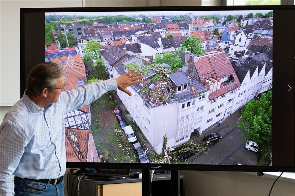 Nach schweren Tornadoschäden: Wüst und Reul besuchen betroffene Gebiete Der Bürgermeister von Paderborn, Michael Dreier, spricht bei einer Pressekonferenz vor einem Display mit einem Schadensbild aus der Innenstadt, das zeigt wie Bäume auf ein Hausdach geweht worden sind.