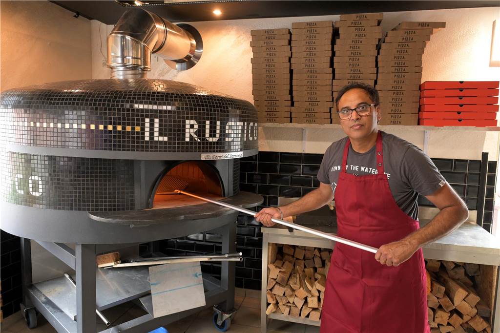 Zwischen Pizza und indischen Speisen: Il Rustico überzeugt im Test Ein Mann mit roter Schürze schiebt eine Pizza in einen schwarzen Holzofen.