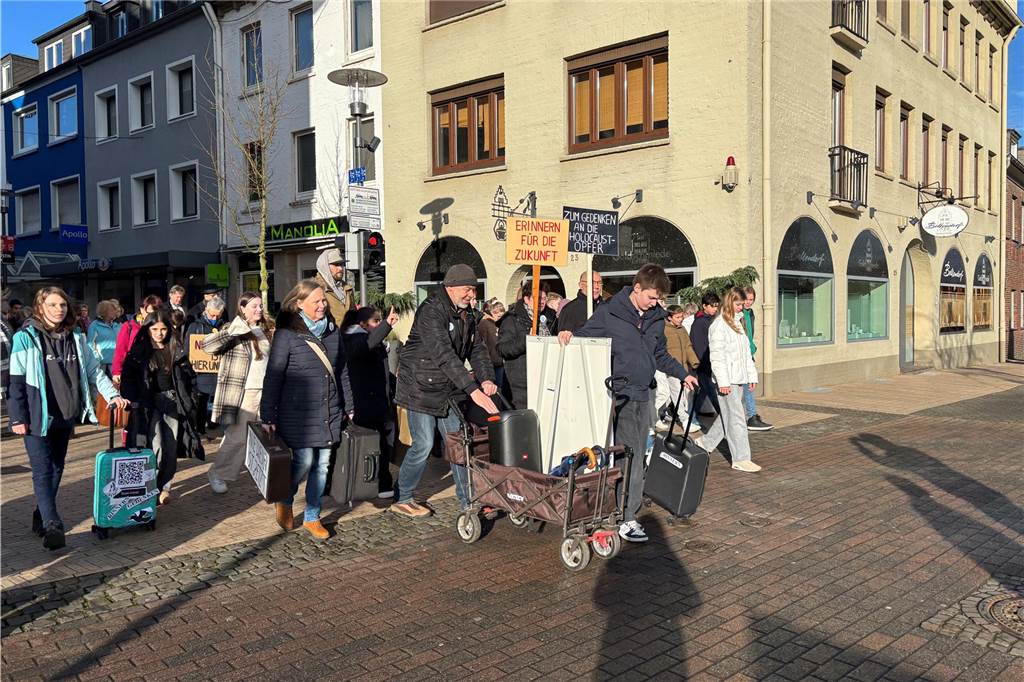Der Koffermarsch ist von vorne fotografiert. Er wird angeführt von Personen mit einem Bollerwagen. Hinter ihnen Personen mit Schildern und Koffern. 