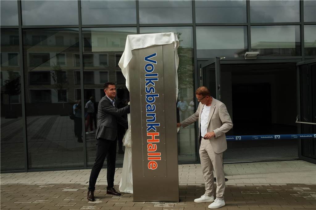 Eröffnung des Burgerladens „Kunstwerk“: Auch Name der Dreifachturnhalle steht Zwei Männer ziehen eine weiße Hülle von einer Säule mit der Gravur „Volksbankhalle”