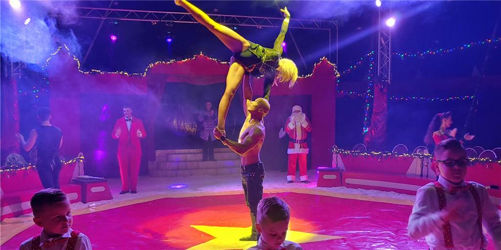 So kann der Weihnachtscircus in Dorsten trotz Energiekrise stattfinden Auf dem Bild sieht man die Zirkusmanege des Weihnachtscircus'. Im Mittelpunkt steht ein Artistenpaar. Der Mann hebt die Frau an einem Fuß und der Hüfte hoch. Im Vordergrund sieht man die nächste Generation der Zirkusfamilie. Im Hintergrund sind weitere Artisten und ein Weihnachtsmann zu sehen.