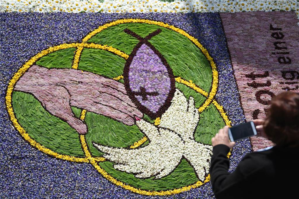 Fronleichnam 2023: Feiertag, Prozession, Bedeutung - alle Infos Eine Frau fotografiert an Fronleichnam während der Prozession Kirchenmotive in einem Blumenteppich.