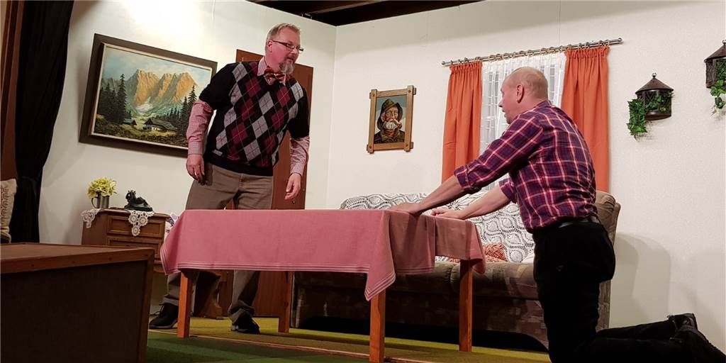 „Corona-Pandemie macht uns Sorgen“: Theatergruppe sagt Aufführung ab Zwei Männer spielen Theater, in der Mitte des Raumes steht ein Tisch.
