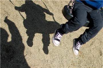 Die Schatten von einem Mann und einem schaukelnden Kind fallen auf einen Spielplatz. Foto: Julian Stratenschulte/Archiv
