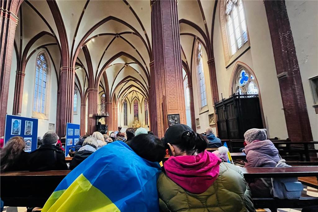 Über 200 Menschen bei Mahnwache zum Ukraine-Krieg in der Sixtuskirche Zwei Menschen sind von hinten in einer Kirche zu sehen. Eine Person hat eine Ukraine-Flagge über den Schultern und lehnt sich mit dem Kopf an der Schulter einer anderen Person an.