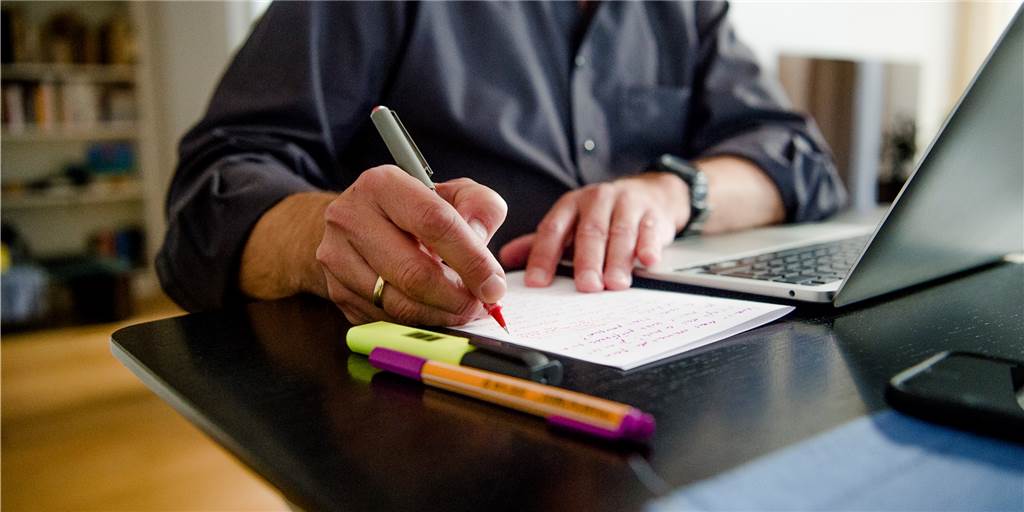 Experiment Vier-Tage-Woche: Mit neuen Konzepten gegen den Fachkräftemangel? Ein Mann sitzt zuhause an einem Esstisch. Er schreibt mit einem Stift auf ein Blatt Papier neben einem Laptop.