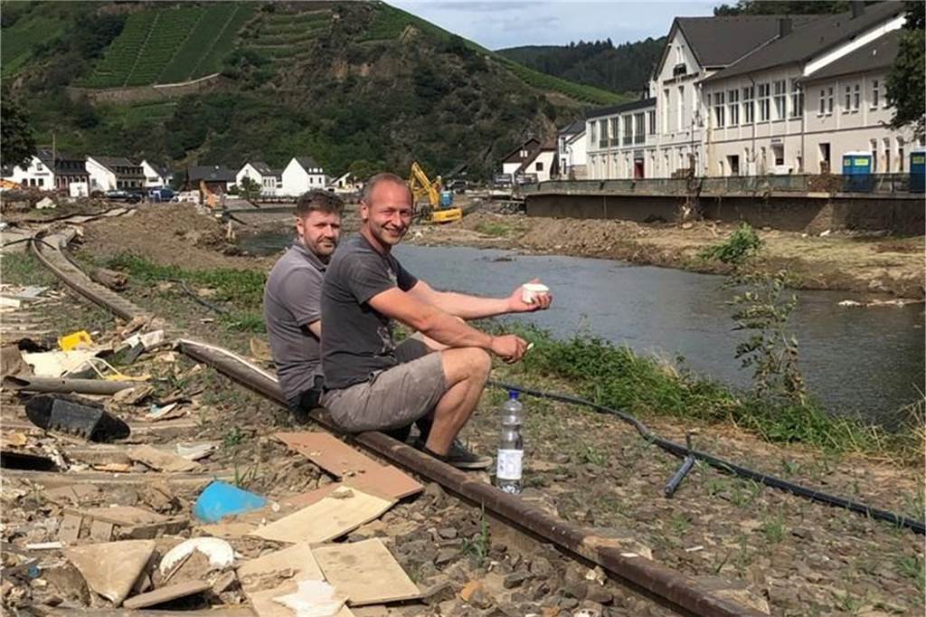 „Unfassbar, dass ein Eis so glücklich machen kann“: Peter Telgmann teilt Eis in Krisenregion aus Dort, wo einmal die Schienen des Bahnverkehrs zu sehen waren, liegen heute Trümmerteile. Dazwischen gönnen sich diese beiden Helfer eine Pause - mit einem „Eis Berger“.