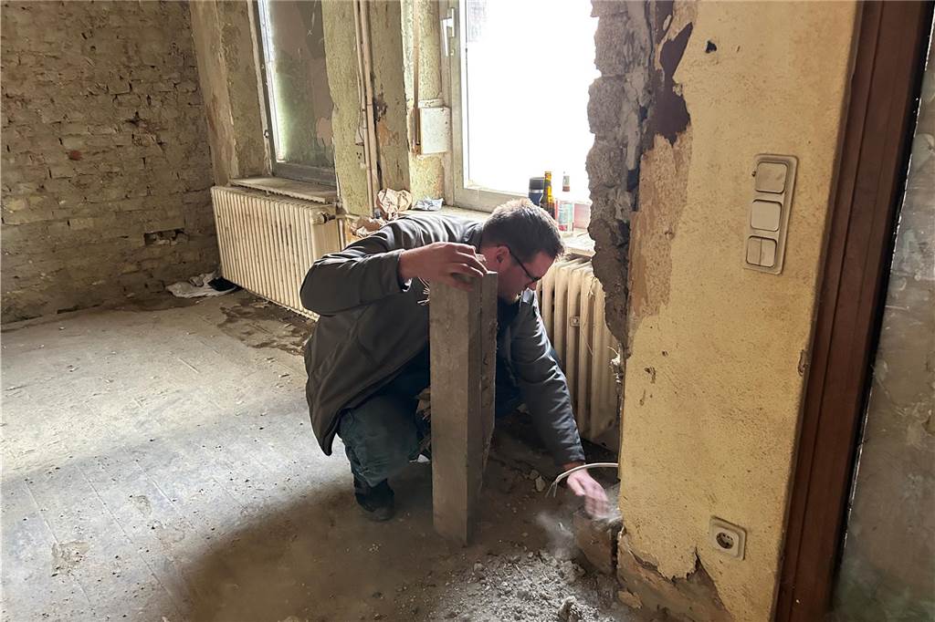 Ein Mann kniet in einem kahlen Raum. An den Wänden sind nur Backsteine. Er prüft ein Holzteil, das von der Wand gebröckelt ist.
