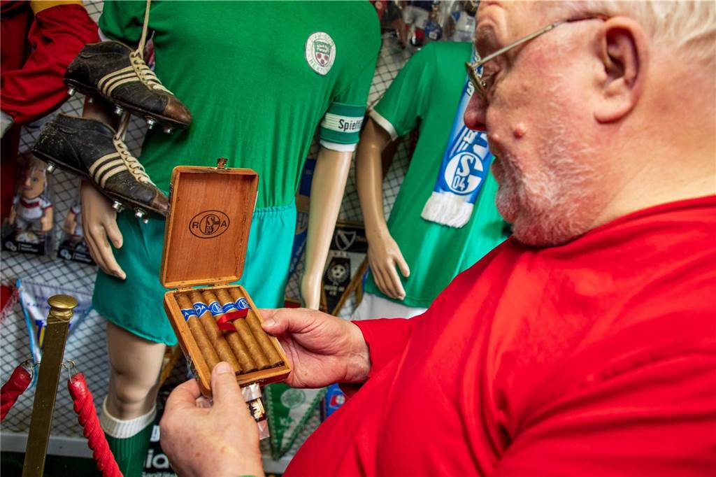 Johann Geiermann (80) hat als Kind mit Rudi Assauer „gepöhlt“ Johann Geiermanns Gesicht ist im Profil zu sehen. Er hält eine hölzerne Schachtel in Händen, in der fünf Zigarren liegen. Jede Zigarre ist mit einer blau-weißen Bordüre versehen, auf der „FC Schalke 04“ und „Rudi Assauer“ eingraviert sind.