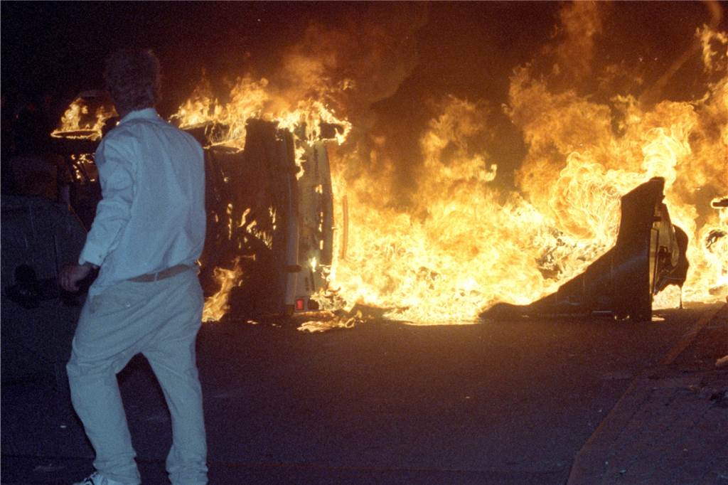 30 Jahre Rostock-Lichtenhagen: „Sowas kann jederzeit wieder passieren, in jeder Stadt“ Ein Mann steht am 27.08.1992 vor einem brennenden Pkw auf einer Straße am zentralen Asylbewerberheim von Mecklenburg-Vorpommern in Rostock-Lichtenhagen.