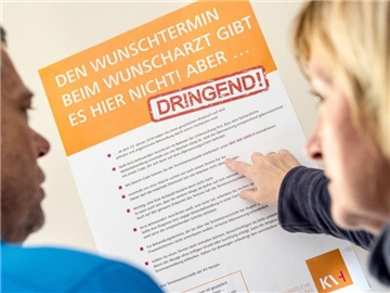 Ein Mann und eine Frau betrachten ein Plakat mit Hinweisen zu der Einführung von "Terminservicestellen". Foto: Alexander Heinl/Archiv