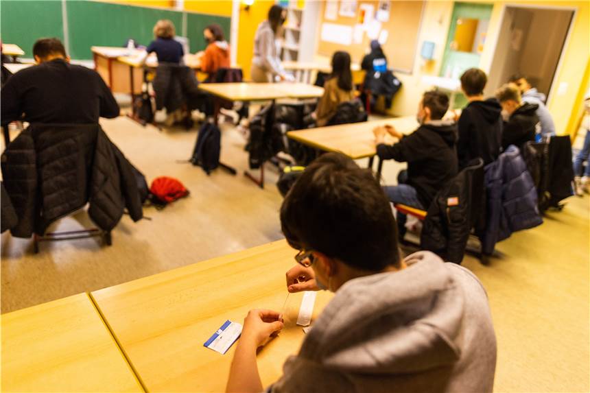 Ein Schüler einer Realschule hält sein Probestäbchen in die Testflüssigkeit für einen Corona-Schnelltest, während im Hintergrund andere Schülerinnen und Schüler zu sehen sind. Hunderte Schüler in NRW lehnen die Tests aber ab und verweigern somit den Präsenzunterricht.