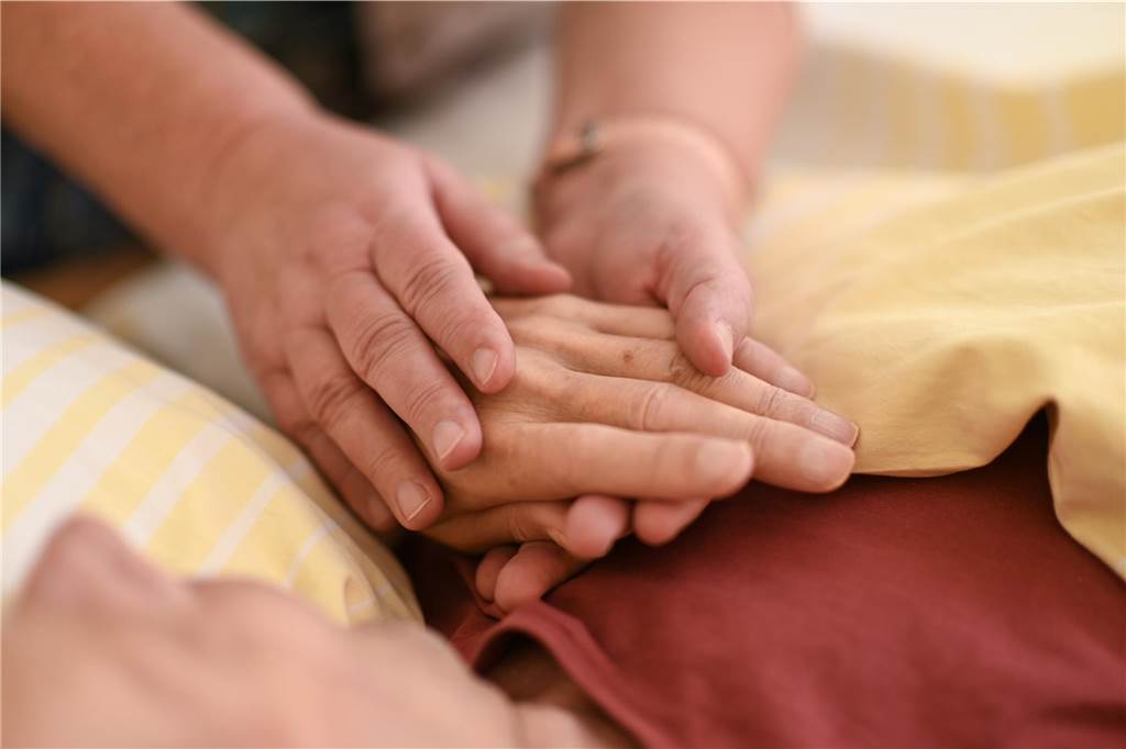 Eine Hospizmitarbeiterin hält die Hand eines todkranken Menschen, der in einem Hospiz im Bett liegt. 