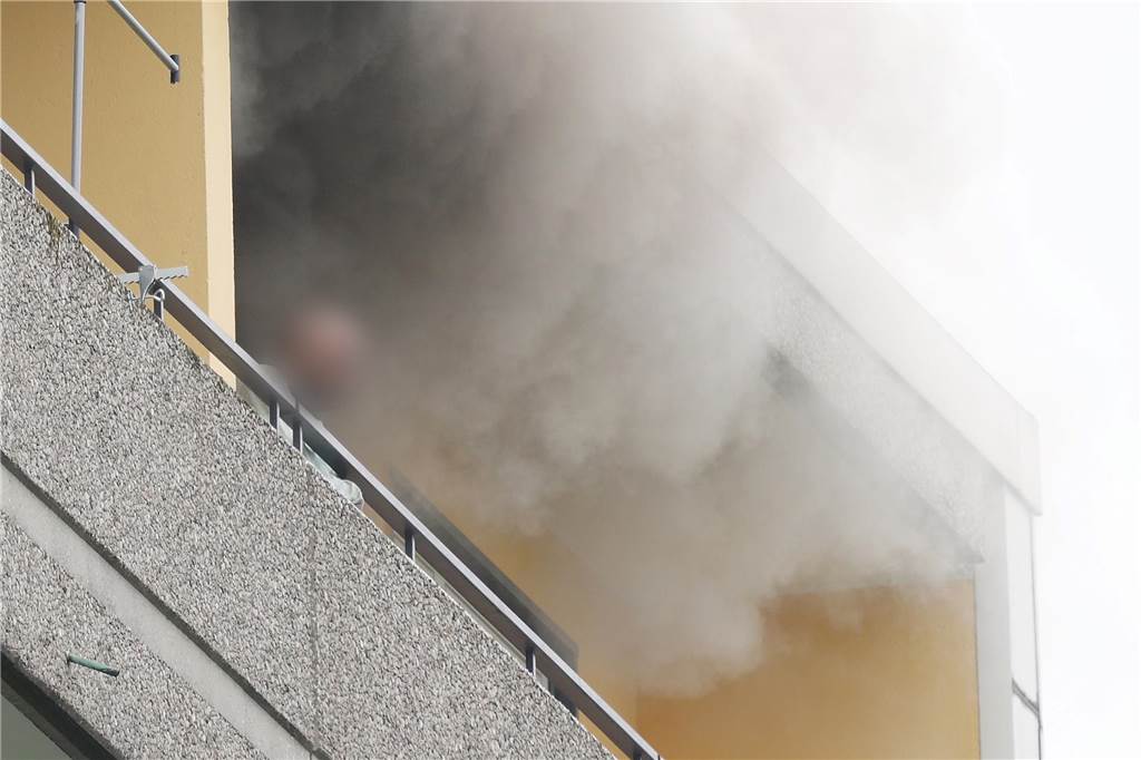 Eine Person steht auf dem Balkon von einem Hochhaus, aus dem Rauch quillt. Bei einer Explosion in einem Hochhaus in Ratingen am Donnerstagvormittag wurden mehrere Menschen verletzt - darunter auch Polizisten und Einsatzkräfte der Feuerwehr. Das erfuhr die Deutsche Presse-Agentur aus Polizeikreisen.