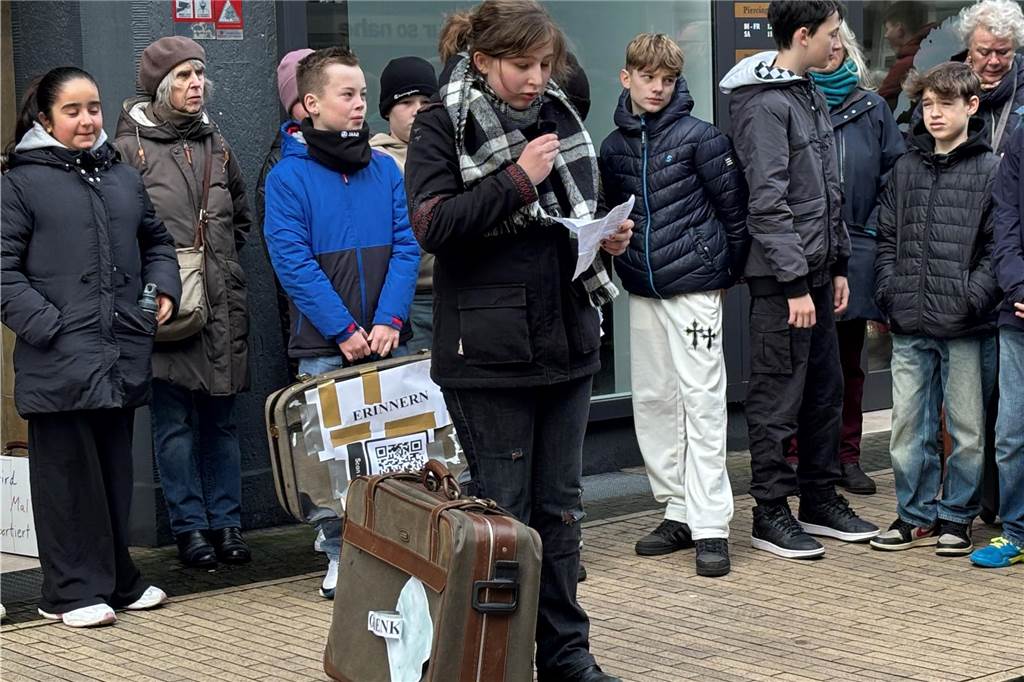 Eine Schülerin hält eine Rede. Neben ihr steht ein alter Koffer. Sie hält ein Mikrofon und liest einen Text vor. Im Hintergrund sind weitere Schüler vor einer Wand zusehen. 