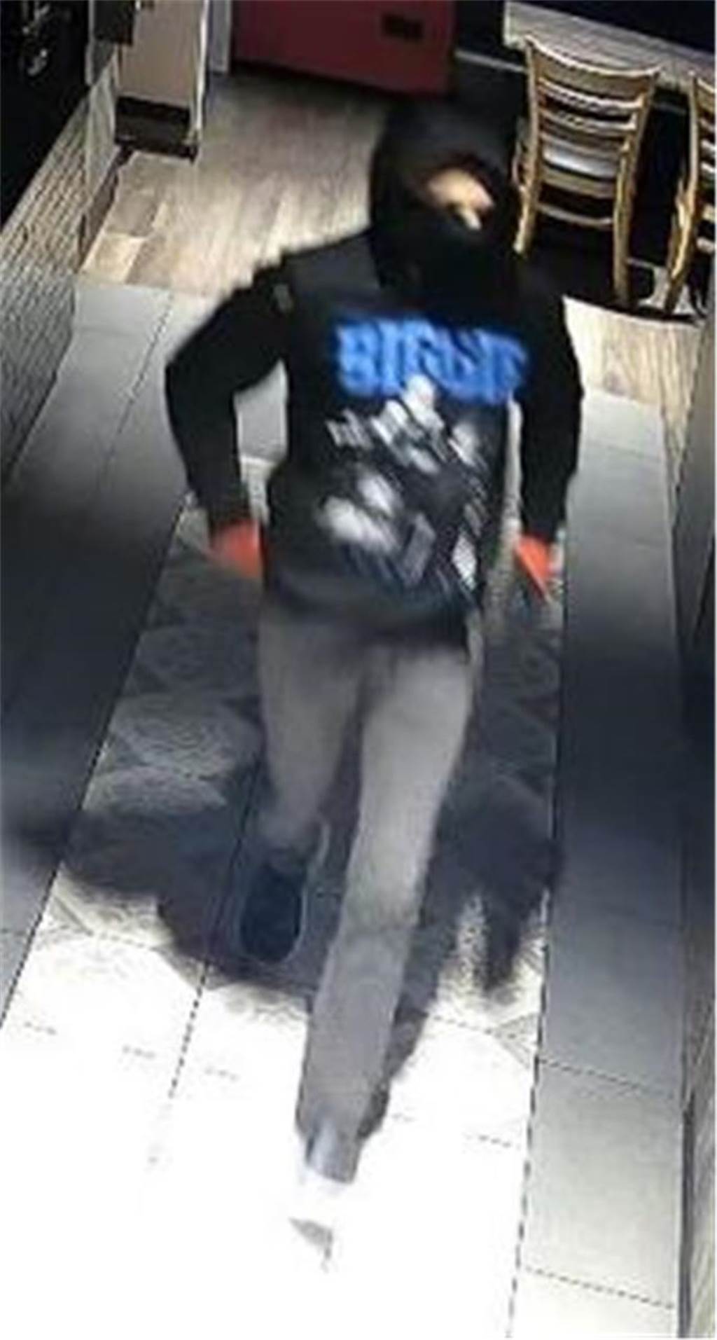 Ein Mann trägt einen Kapuzenpullover mit Aufschrift „Biggie“ und Aufdruck von geringfügig erkennbaren Personen, augenscheinlich Tuch oder Mundmaske aufgezogen, orange-schwarzfarbene Handschuhe, dunkelgraue Jeanshose und dunkle sportliche Schuhe
