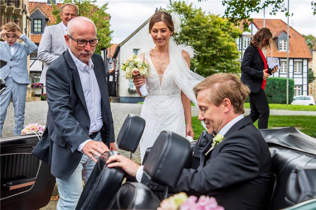 Celine Gallwas und Markus Hilfert geben sich das Jawort Markus Hilfert sitzt auf dem Rücksitz eines Cabrios. Fred Toplak hält eine Hand an einem Sitz, während Celine Gallwas am Auto steht und mit dem Brautstrauß in der Hand zuschaut.