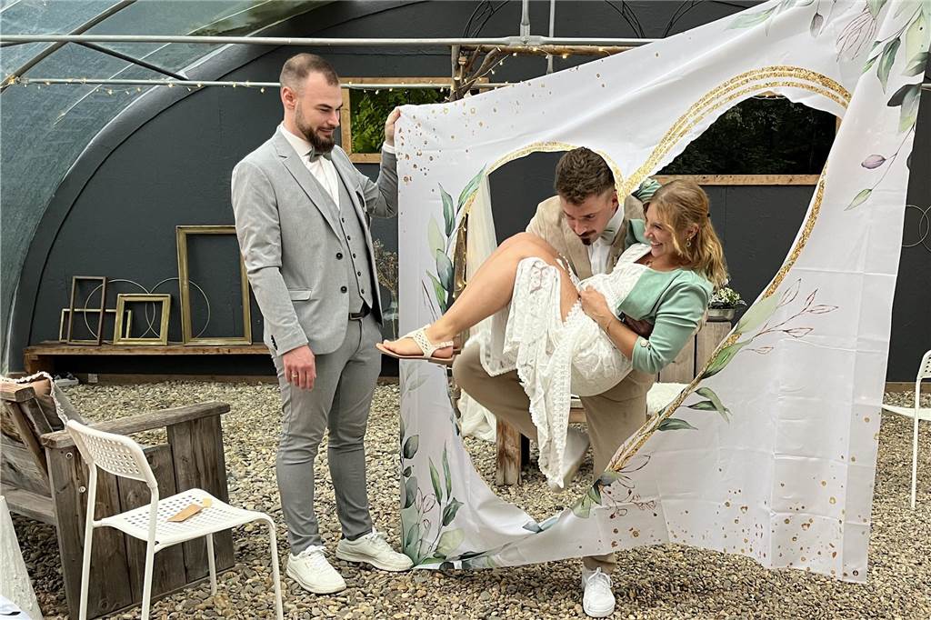Ein Hochzeitsgast hält ein Tuch, in das ein Herz geschnitten wurde. Bräutigam Michael trägt seine Braut Elena durch das Herz-Loch.