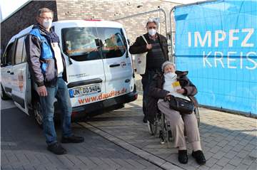 Fahrer Karl-Heinz Hug mit Klaus und Margret Bekemeier nach ihrem Impftermin vor dem himmelblauen Transparent des Impfzentrums Unna: Beide sind dankbar für die Hilfestellung durch den AWO-Fahrdienst.