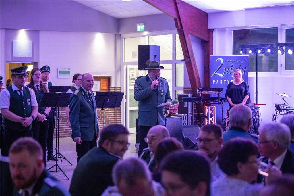 Vor dem Spielmannszug stehen die Vereinsvorsitzenden und begrüßen die Gäste. Im Vordergrund sitzen Menschen.