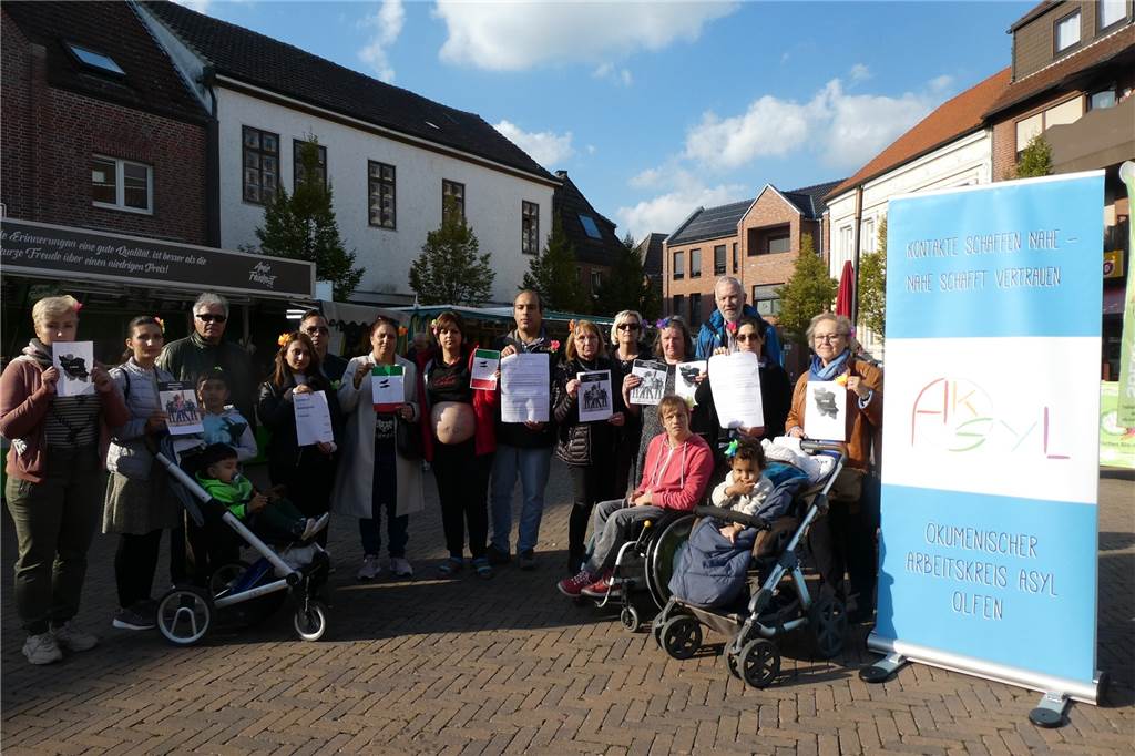 Eine Gruppe Menschen steht auf dem Marktplatz in Olfen neben einem großen Plakat des Arbeitskreises Asyl. 