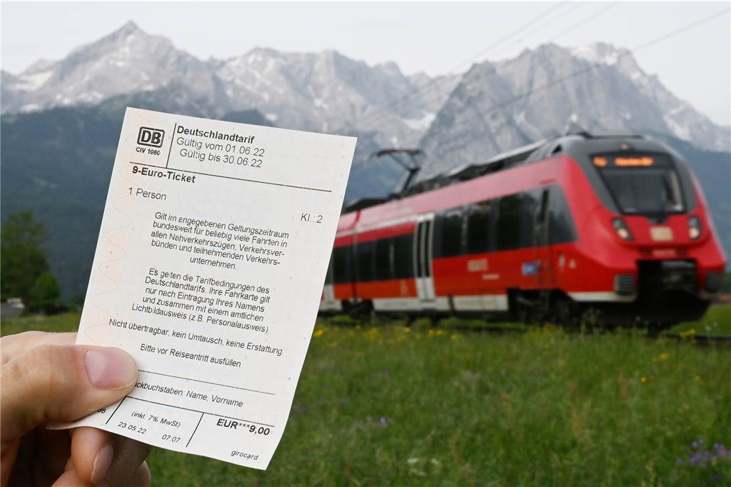 Wann Reisende mit dem 9-Euro-Ticket auch Fernverkehrszüge nutzen dürfen Garmisch-Partenkirchen: Ein Mann hält ein 9-Euro-Ticket in der Hand, im Hintergrund fährt ein Zug der Deutschen Bahn vor der Bergkulisse des Wettersteingebirges vorbei.