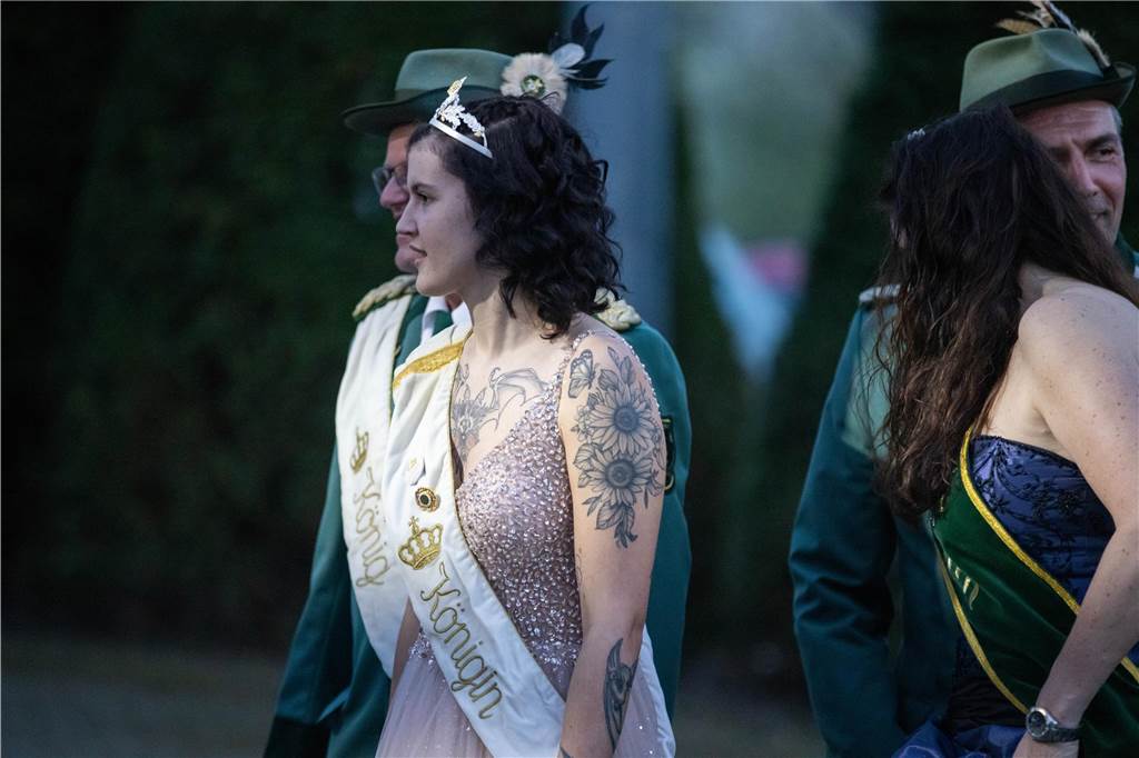 Eine Frau mit dunklen Locken und Tattoos auf Brust und Oberarmen trägt ein Diadem, ein Rosanes Kleid und eine weiße Scherpe mit der Aufschrift: Königin.
