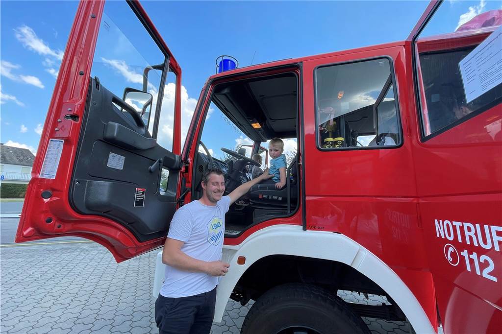 Ein Junge sitzt auf dem Fahrersitz eines Feuerwehrautos, sein Vater steht davor und passt auf.