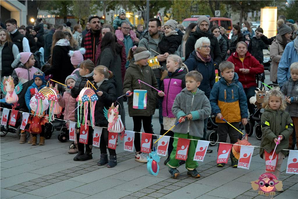 Viel Kinder mit Laternen stehen hinter einem Absperrband und halten Laternen in der Hand. Hinter ihnen stehen Erwachsene. 